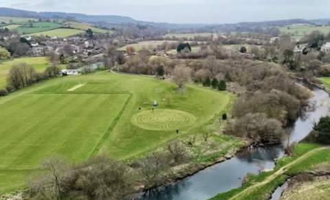 Aerial view of a countryside landscape featuring a sports field with goalposts, a circular grassy labyrinth, and a winding river. Rolling hills and scattered trees convey a peaceful, rural vibe.