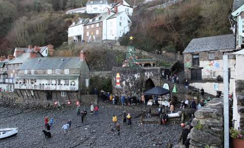 Coastal village scene with festive decorations. People gather near stone buildings and a Christmas tree. Pebbled shore in foreground and hillside houses behind.