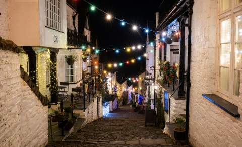 A charming cobblestone street at night, lined with cosy houses adorned with glowing multicoloured string lights, creating a festive, warm atmosphere.