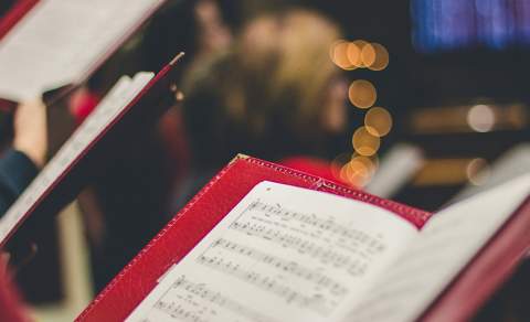 Close-up of sheet music in red folders held by choir singers, blurred background with warm bokeh lights, conveying a festive and serene atmosphere.