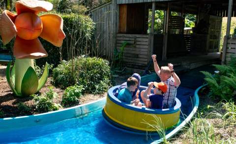 A group of people enjoy a log flume ride, passing by a large, whimsical orange flower sculpture. Lush greenery surrounds the area.