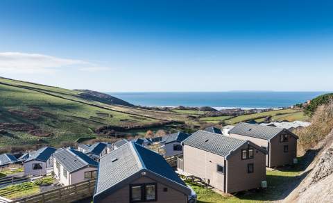 Scenic view of a hillside with modern cabins overlooking the ocean. Green fields stretch toward the sea under a clear blue sky, conveying tranquillity.