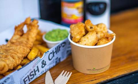 A hearty serving of fish and chips with mushy peas in a newspaper-lined box on a wooden table. A paper cup with fried snacks is nearby, evoking a warm, casual dining experience.