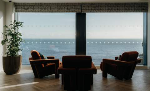 A tranquil room with three plush chairs facing a large window, offering a serene ocean view. A potted plant adds a touch of greenery to the cosy space.