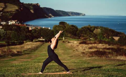 A person practices yoga outdoors on a grassy path with arms reaching up. The backdrop shows cliffs, a coastal village, and a calm blue sea.