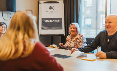 A small group of people and assistance dog sit around a meeting table in a conference room, engaged in discussion. A flip chart at the front of the room displays the DoubleTree by Hilton logo with the word “Welcome.” Natural light comes through a large window, and notebooks and papers are spread across the table.