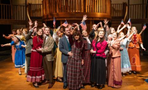 NAPA cast stands on stage, they are dressed in American colonian costumes, smiling and waving American flags.