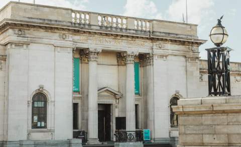 Front exterior of the Ferens Art Gallery in Hull, showing its neoclassical stone façade with tall columns, decorative carvings and entrance banners on a bright day, with the nearby historic lamp feature in the foreground.