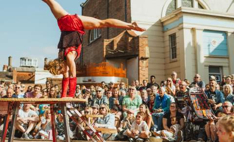 A woman in a red leotard is performing a hand stand on a raised platform with a crowd surrounding her.