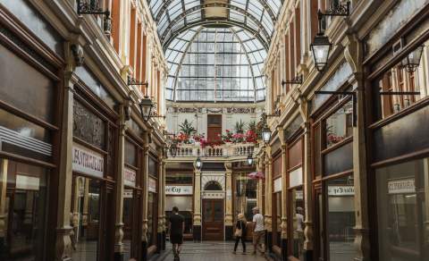 Hepworth Arcade in the summer sunshine. The arched windows show the tower of Hull Minster in the background. Below, the lovingly tendered plants and flowers sit on the windowsill. Shoppers stroll the shops and Victorian lamps line the doorways.