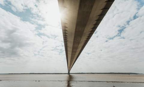 A view beneath the Humber bridge, with clouds either side and the foreshore just in front of the viewer.