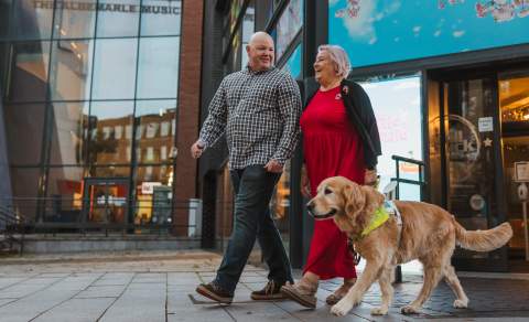 A man and woman walking with a guide dog along a city street outside Hull Truck Theatre.