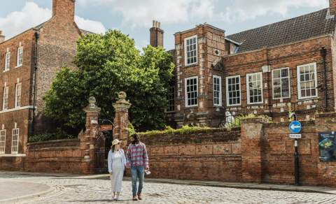 A cobbled street scene outside a historic red‑brick building in Hull’s Museums Quarter, with two visitors walking past the Wilberfoce House under a bright, clear sky.