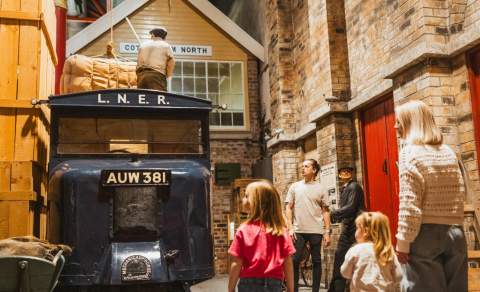 Families exploring a historic railway-themed gallery, looking at an L.N.E.R. vintage delivery truck, luggage display, and recreated station setting inside a Streetlife Museum Hull .