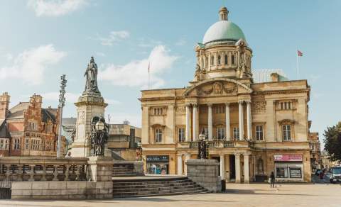 Queen Victoria Square on a sunny day. Hull City Hall is in the background, the early 20th-century Baroque Revival columns and blue copper dome look striking in the sun, The statue of Queen Victora is standing proud to the left of the image.