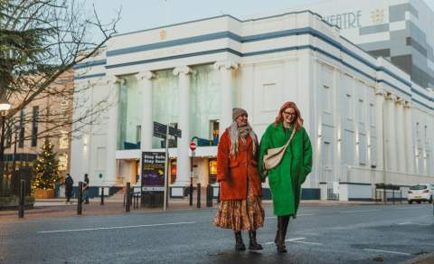 Hull New Theatre in the winter, the white and blue building is light by vertical lights up the colums. Two ladies are walking away after seeing a show. They are bundled up in hats and coats. The lady on the left has blonde hair and is wearing a rusy red fur coat and a beige hat. The lady on the right is wearing a green coat and has red hai and glasses.