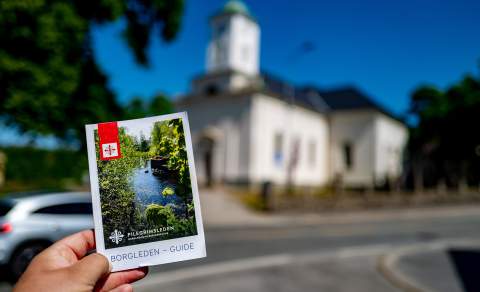 En hånd holder i en brosjyre for Borgleden foran en kirke