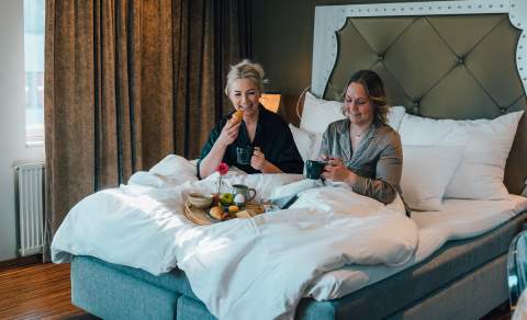 To damer som spise mat og drikke kaffe i en hotellseng. De ler og hygger seg.