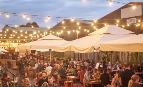 Outdoor dining area with long tables, string lights above, and large umbrellas. People are enjoying food and drinks at dusk, creating a lively and warm atmosphere.