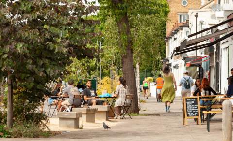 A lively street scene with people dining outdoors at café tables and benches, surrounded by greenery and trees. Evoking a relaxed, community vibe.
