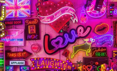 A vibrant collage of colorful neon signs in various shapes and words, including "Love," "Laughter," and "Applause." At God's Own Junkyard in Walthamstow, the scene is bright and lively.