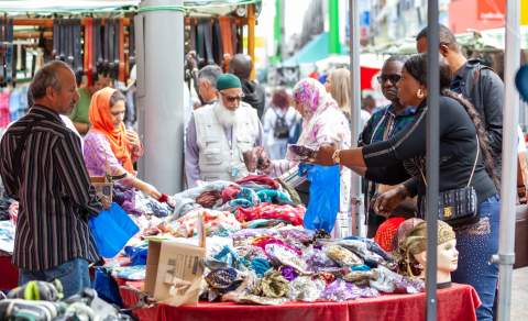 A bustling outdoor market scene with diverse shoppers examining colourful fabrics on a table. Mannequin heads display headscarves, creating a lively, multicultural atmosphere.