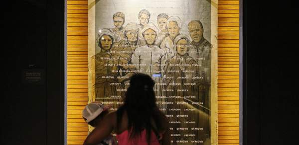 A Black woman with long hair holds her child while they look at a photo collage of formerly enslaved residents of Africatown, Alabama