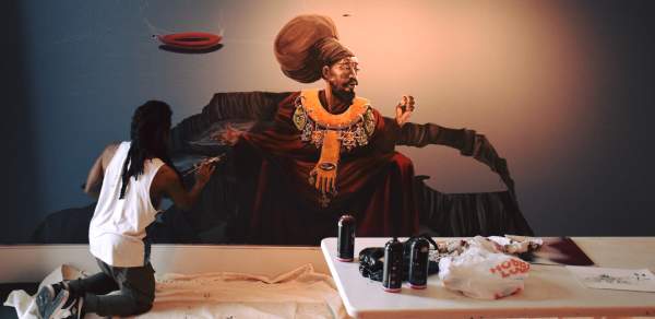 An artist kneels as he paints an African woman on a wall with artist supplies on a table