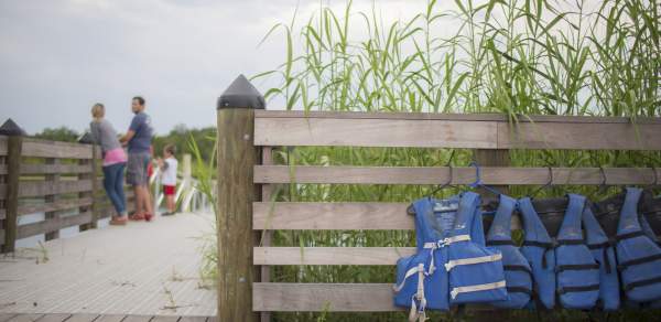 people walking down the boardwalk to the beach, fence with life jackets hanging
