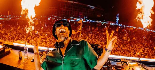 DJ Fisher performs on the stage in front of a crowd with flames in the background.