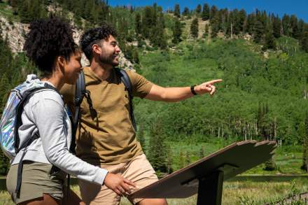 Man and Women with day hike backpacks looking off screen and pointing with a scenic background and information sign in front of them