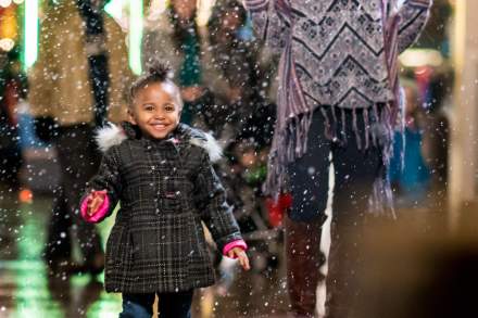 A mom and daughter with decorative holiday lights behind them look with anticipation toward a fancy chair