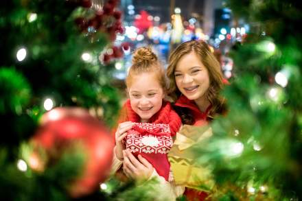 Looking at two happy girls through the branches of an evergreen with lights on it