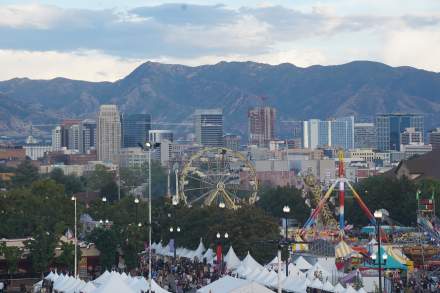 Aerial view of fair tents and carnival rides including a ferris wheel with the Salt Lake skyline and Wasatch Mountains in the background
