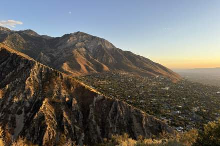 Edge of one mountain with another mountain in the background leading down to a populated area  of the Salt Lake valley with trees and houses