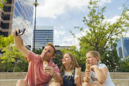 Father taking a selfie with 2 daughters all eating ice cream. In the background are the buildings of downtown Salt Lake