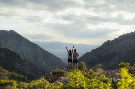 Couple standing on a rock with arms up the view looks down through a canyon into the Salt Lake valley