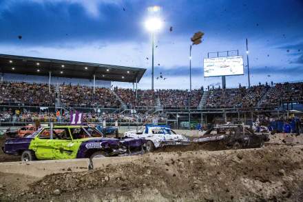 Demolition derby cars crashing into each other with mud flying and filled stadium seats behind them.