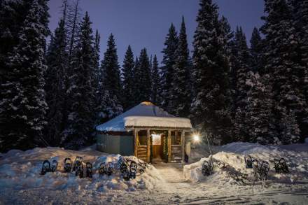 A yurt lit up by lanterns on each side of the door in the snow with large pines in the background