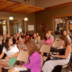 A group of people sitting down, looking towards one woman who is standing and speaking