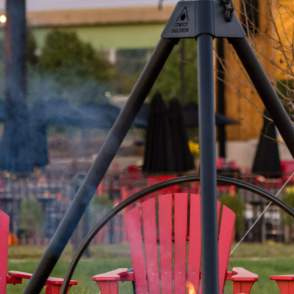 People sitting around a fire in red Adirondack chairs, in the back there is a building and an outdoor stage