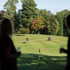A golfing green with trees in the background and people holding drinks in the foreground