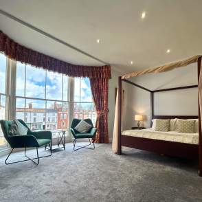 a bed and two chairs in a hotel room by a bay window overlooking a town square