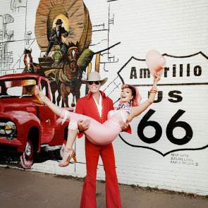 A man dressed in a red Western-inspired suit with flared pants, a cowboy hat, sunglasses, and blue shoes is holding a woman in a pink vintage-style outfit. The woman is playfully posing with one leg bent, wearing high heels, a scarf around her neck, and holding a pink cowboy hat in the air. They are both smiling in front of a mural depicting a classic red truck, a covered wagon with a cowboy, and the words "Amarillo US 66" painted on a white brick wall. The image captures a lively, retro, and playful Route 66-themed moment.
