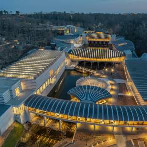 Aerial view of a modern architectural complex with flowing lines, illuminated pathways, and surrounding greenery at dusk. The design features multiple interconnected buildings and water elements.