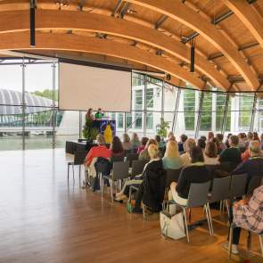 People sitting in a Crystal Bridges Museum of American Art great hall. The room has glass windows and wooden beams on the ceiling.