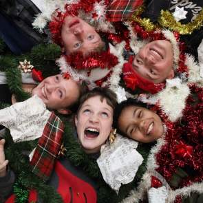 Photo of the upcoming theatre production of The Best Christmas Pageant Ever. 5 Kids are laying down in a circle, heads first and facing up, with christmas decorations all around them.
