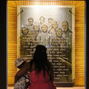 A Black woman with long hair holds her child while they look at a photo collage of formerly enslaved residents of Africatown, Alabama