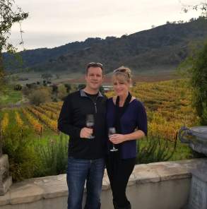 Couple standing together with wine glasses in front of vineyard scenery during harvest season.