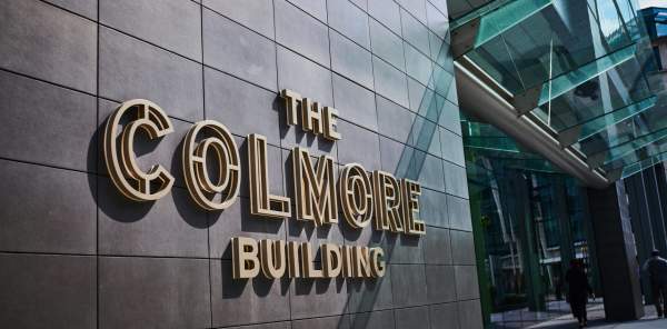 Raised golden signage lettering reads THE COLMORE BUILDING next to the building entrance under a glass canopy